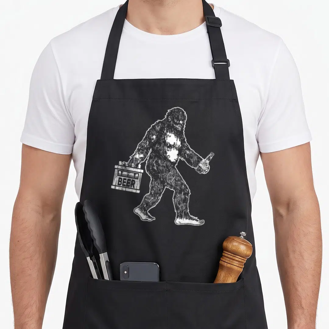 Bigfoot craft apron - White line-art bigfoot holding beer carrier and bottle — shown on a black cotton canvas apron against a white background