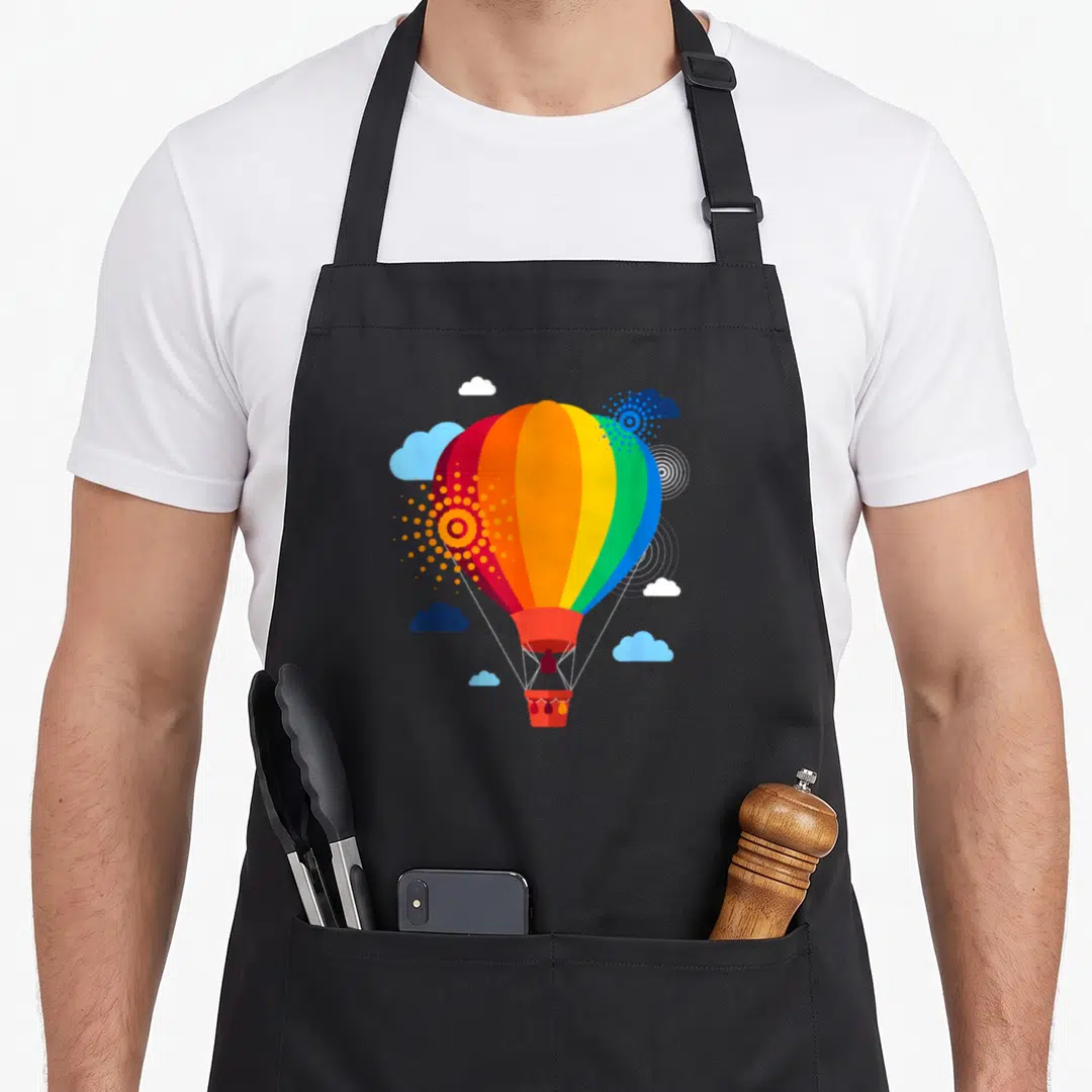 Ride balloon apron - Cartoon hot air balloon with red, orange, yellow, green, and blue stripes surrounded by scattered — shown on a black cotton canvas apron against a white background