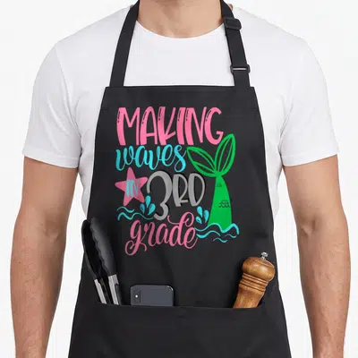 Mermaid art teacher aprons - Making waves in 3rd grade text with green mermaid tail and pink star — shown on a black cotton canvas apron against a white background