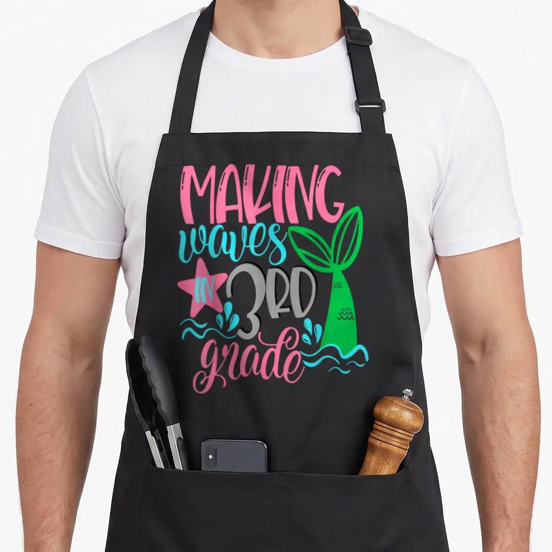 Mermaid art teacher aprons - Making waves in 3rd grade text with green mermaid tail and pink star — shown on a black cotton canvas apron against a white background