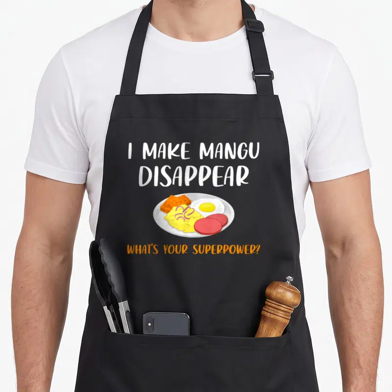 Superpower pride apron - Cartoon plate of mangu, sausage, egg, tostones with I Make Mangu Disappear text — shown on a black cotton canvas apron against a white background