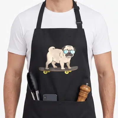 Skateboarding dog apron - Cartoon beige pug with blue sunglasses standing on a grey skateboard with yellow wheels — shown on a black cotton canvas apron against a white background