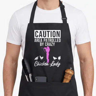 Caution chicken apron - Pink woman silhouette with binoculars surrounded by four white chickens and caution text — shown on a black cotton canvas apron against a white background
