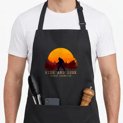 Cool hide bigfoot apron - Bigfoot silhouette against orange sun and red trees with Hide and Seek World Champion text — shown on a black cotton canvas apron against a white background