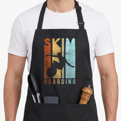 Skim surfer apron - Grey skimboarder silhouette over orange, yellow, and teal vertical stripes with SKIM BOARDING text — shown on a black cotton canvas apron against a white background