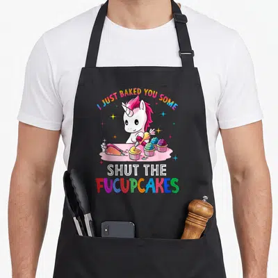 Baked shut the fucupcakes apron - Cartoon unicorn piping rainbow frosting with I Just Baked You Some Shut — shown on a black cotton canvas apron against a white background
