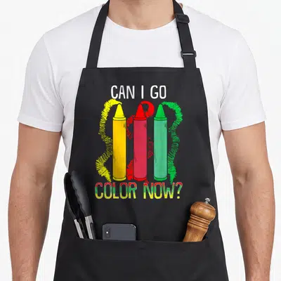 Crayon classroom aprons - Can I Go Color Now text with three vertical yellow red and green crayons — shown on a black cotton canvas apron against a white background