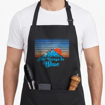 Player bluegrass apron - Retro mountain landscape with blue orange red stripes and My Grass Is Blue text — shown on a black cotton canvas apron against a white background
