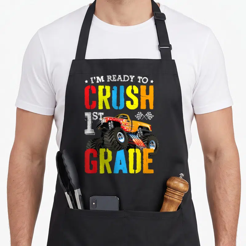 1st truck apron - I'M READY TO CRUSH 1ST GRADE text surrounding a cartoon monster truck with checkered flags and stars — shown on a black cotton canvas apron against a white background