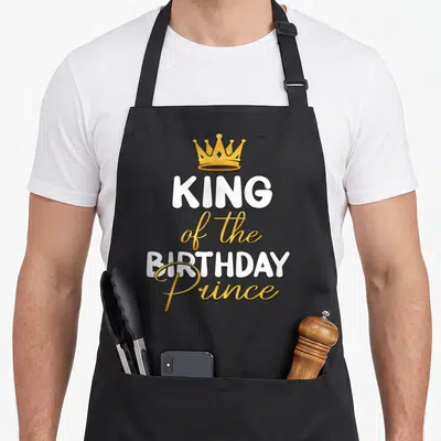 Funny birthday king apron - Gold crown above white and gold text reading KING of the BIRTHDAY Prince — shown on a black cotton canvas apron against a white background