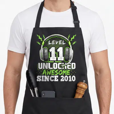 Level unlocked 11th birthday video game apron - Gaming headset illustration with Level 11 Unlocked text and lightning — shown on a black cotton canvas apron against a white background