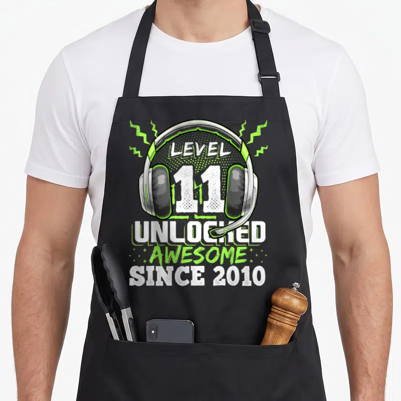 Level unlocked 11th birthday video game apron - Gaming headset illustration with Level 11 Unlocked text and lightning — shown on a black cotton canvas apron against a white background