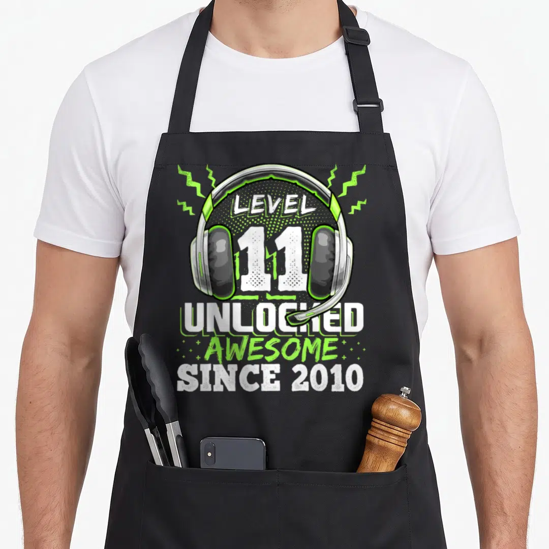 Level unlocked 11th birthday video game apron - Gaming headset illustration with Level 11 Unlocked text and lightning — shown on a black cotton canvas apron against a white background