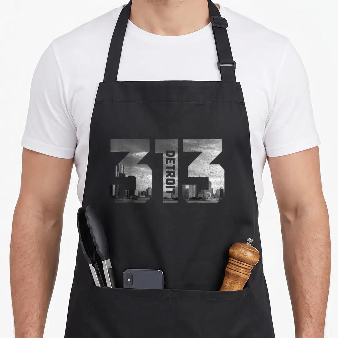 Michigan area pride apron - 313 numbers filled with Detroit skyline photo and vertical DETROIT text in grey and black — shown on a black cotton canvas apron against a white background