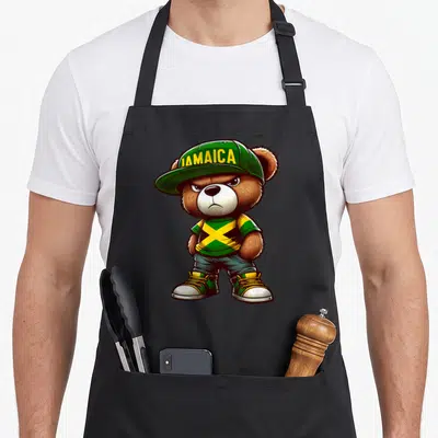 Jamaica teddy bear apron - Cartoon bear wearing green cap with JAMAICA text and flag pattern shirt — shown on a black cotton canvas apron against a white background