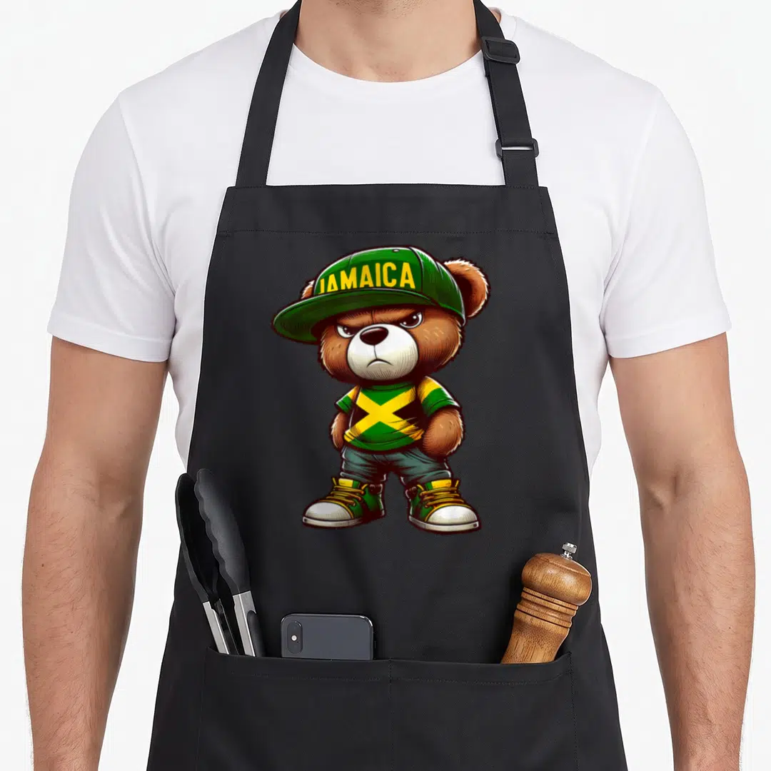 Jamaica teddy bear apron - Cartoon bear wearing green cap with JAMAICA text and flag pattern shirt — shown on a black cotton canvas apron against a white background