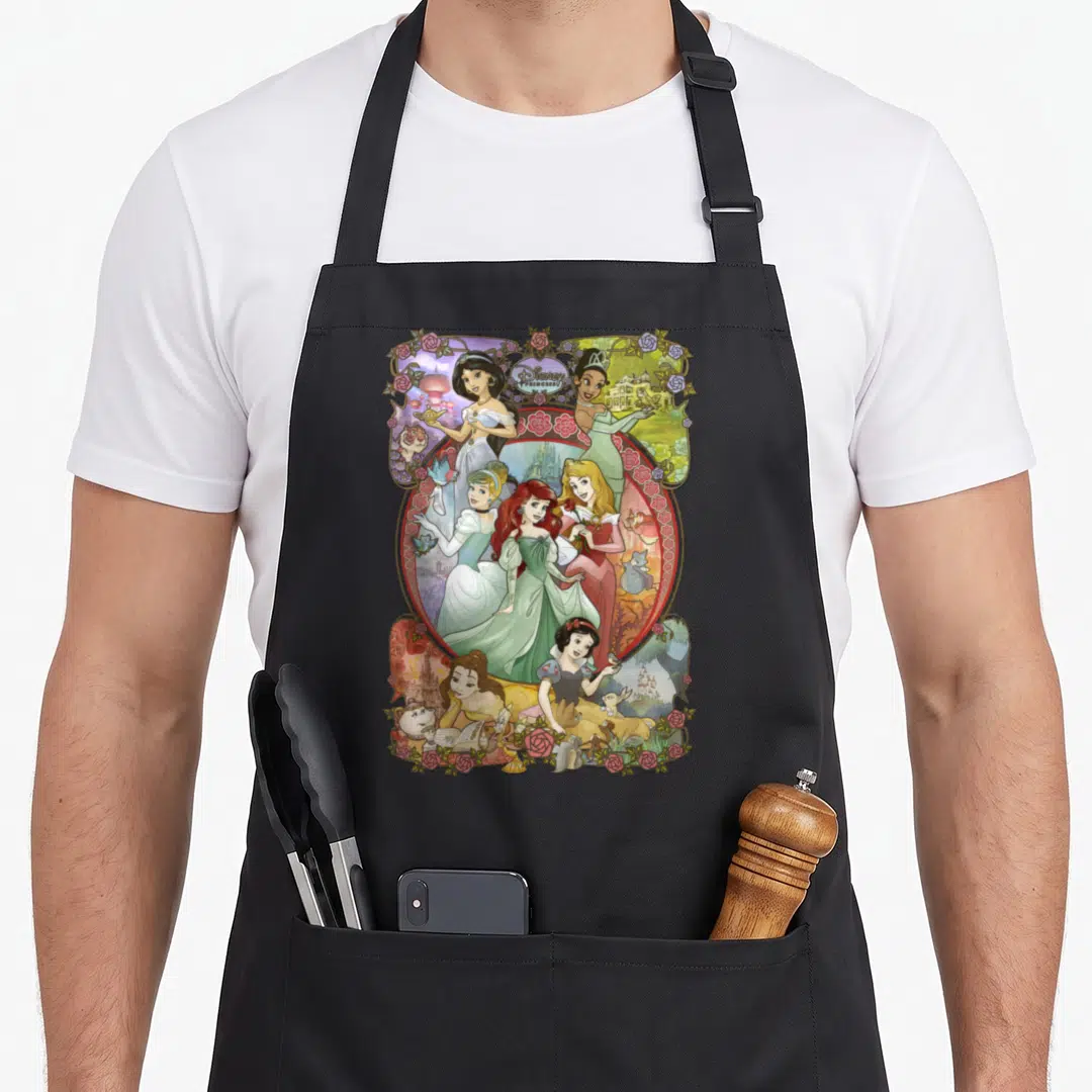 Cartoon princess apron - Six animated princesses in an oval frame surrounded by pink roses and vines with Disney text — shown on a black cotton canvas apron against a white background
