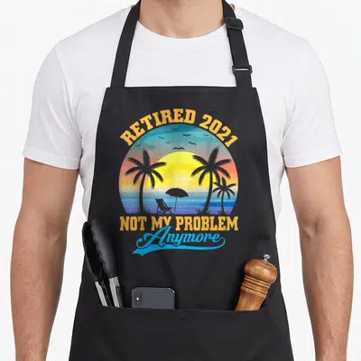 Cute problem retirement apron - Retro beach sunset circle with palm trees, chair, and text Retired 2021 Not My Problem — shown on a black cotton canvas apron against a white background
