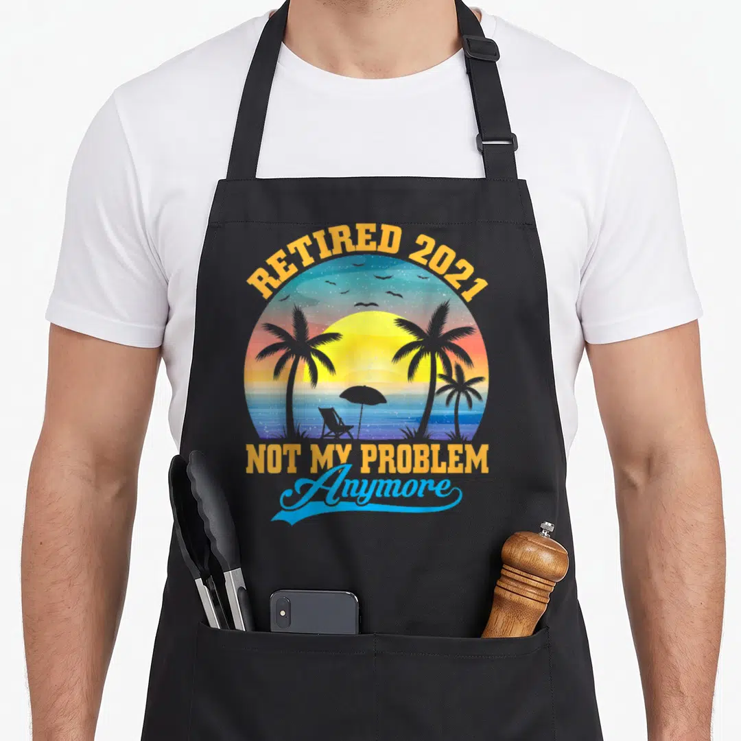 Cute problem retirement apron - Retro beach sunset circle with palm trees, chair, and text Retired 2021 Not My Problem — shown on a black cotton canvas apron against a white background