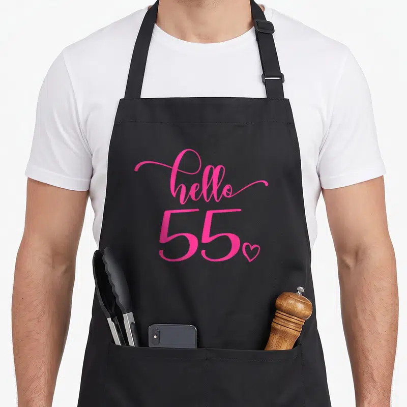 55th fifty birthday apron - Pink hello 55 script text with a small heart outline below — shown on a black cotton canvas apron against a white background