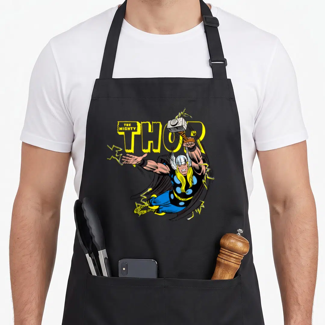 Vintage comic hobby apron - Cartoon Thor flying with hammer and lightning, yellow text THE MIGHTY THOR — shown on a black cotton canvas apron against a white background