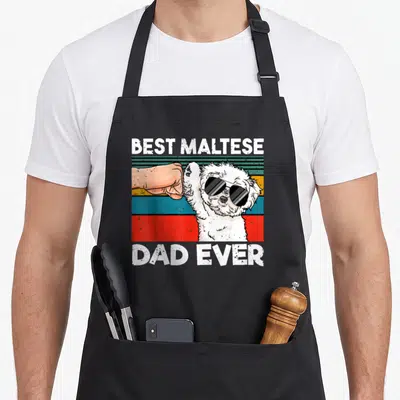 Ghetto dog apron - White Maltese dog in sunglasses fist-bumping human hand under BEST MALTESE DAD EVER text on striped — shown on a black cotton canvas apron against a white background