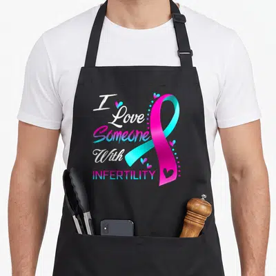 Someone infertility awareness apron - I Love Someone With INFERTILITY text with pink-blue ribbon and small hearts — shown on a black cotton canvas apron against a white background