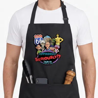 Jeff dunham tulsa apron - Route 66 sign, Jeff Dunham puppets, gold statue, neon SERIOUSLY text, and building background — shown on a black cotton canvas apron against a white background