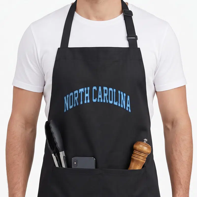 North simple apron - Arched blue NORTH CAROLINA text in retro varsity font — shown on a black cotton canvas apron against a white background