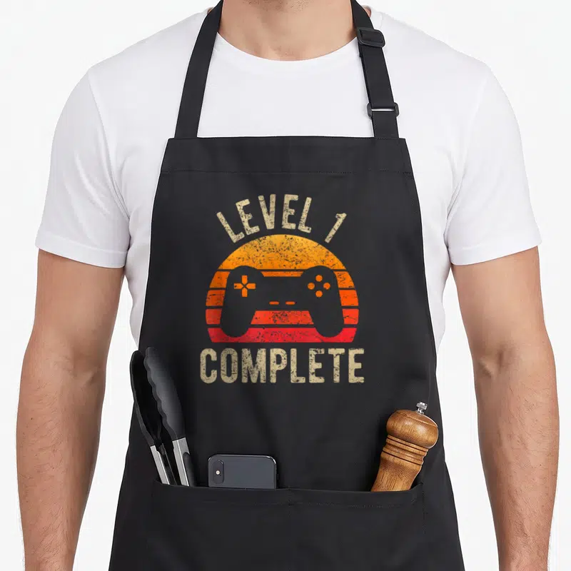 1st wedding apron - Retro striped sun behind grey game controller with beige Level 1 Complete text — shown on a black cotton canvas apron against a white background