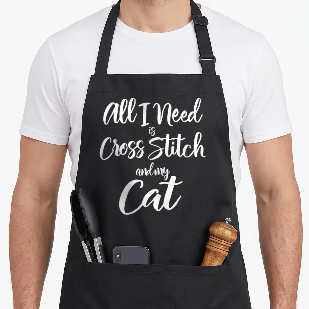 Cat cross stitch aprons - White cursive text reading All I Need is Cross Stitch and my Cat on black fabric — shown on a black cotton canvas apron against a white background