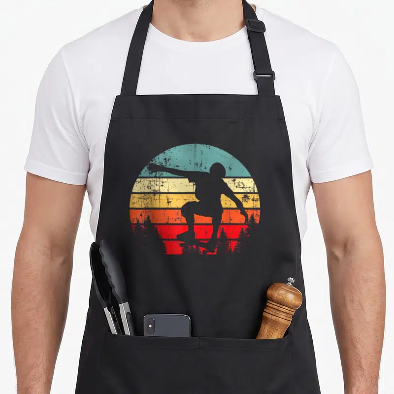 Trick skateboarder apron - Black skater silhouette against retro sun with teal yellow orange red and black stripes — shown on a black cotton canvas apron against a white background