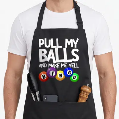 Pull bingo apron - White text PULL MY BALLS AND MAKE ME YELL above five colorful BINGO balls — shown on a black cotton canvas apron against a white background