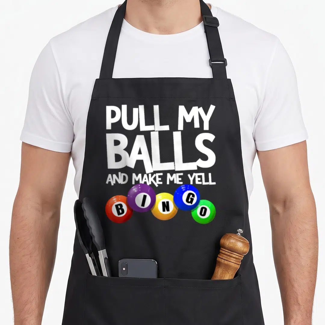 Pull bingo apron - White text PULL MY BALLS AND MAKE ME YELL above five colorful BINGO balls — shown on a black cotton canvas apron against a white background