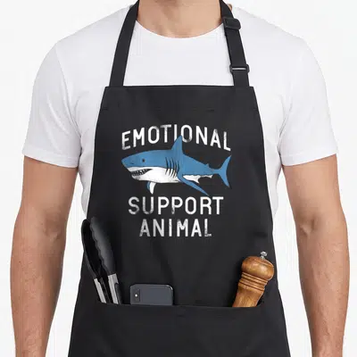 Emotional animal apron - Blue cartoon shark illustration with white text EMOTIONAL SUPPORT ANIMAL on black background — shown on a black cotton canvas apron against a white background