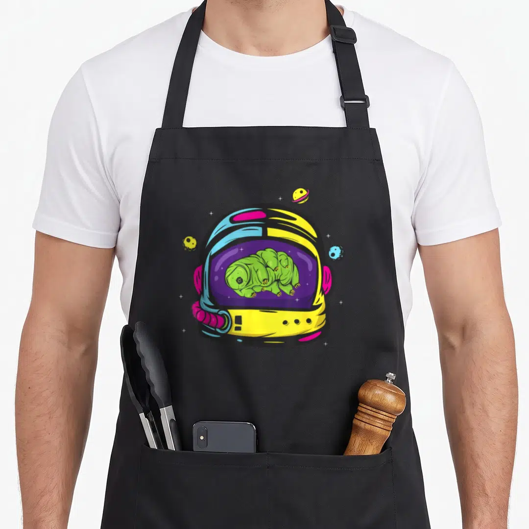 Space bear apron - Green tardigrade curled in yellow blue astronaut helmet with three planets and stars — shown on a black cotton canvas apron against a white background