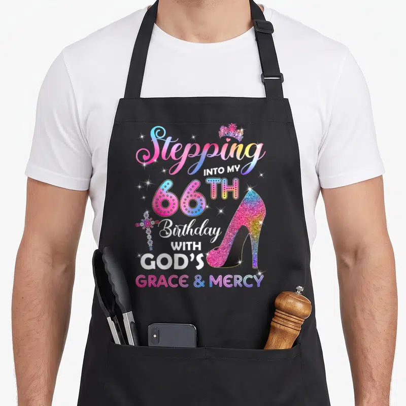 Stepping 66th birthday apron - Glittery pink high heel shoe next to stacked text Stepping Into My 66th Birthday — shown on a black cotton canvas apron against a white background