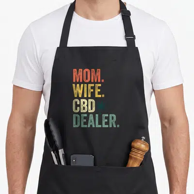 Cbd wife apron - Stacked MOM WIFE CBD DEALER text with cannabis leaf and droplet in orange yellow beige green — shown on a black cotton canvas apron against a white background