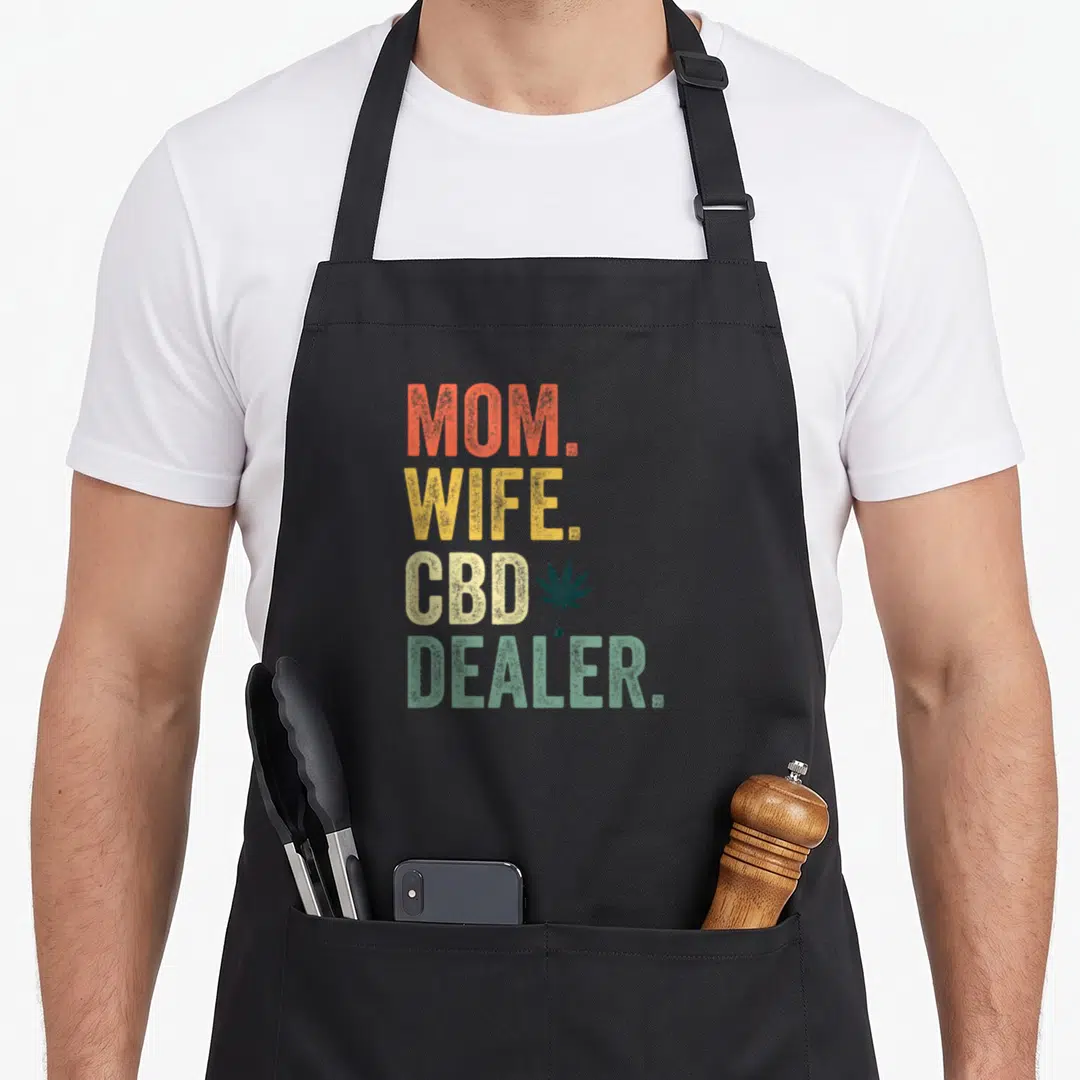 Cbd wife apron - Stacked MOM WIFE CBD DEALER text with cannabis leaf and droplet in orange yellow beige green — shown on a black cotton canvas apron against a white background