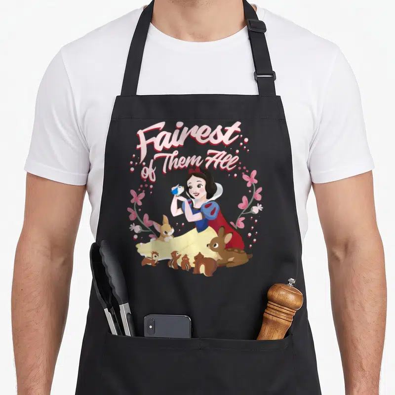 Fairest floral apron - Snow White with bird and animals inside pink flower wreath under arched Fairest of Them All text — shown on a black cotton canvas apron against a white background