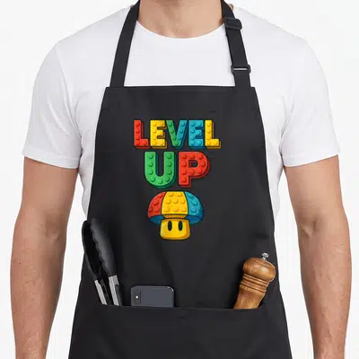 Level block & apron - LEVEL UP text and brick-style mushroom in red yellow blue green — shown on a black cotton canvas apron against a white background
