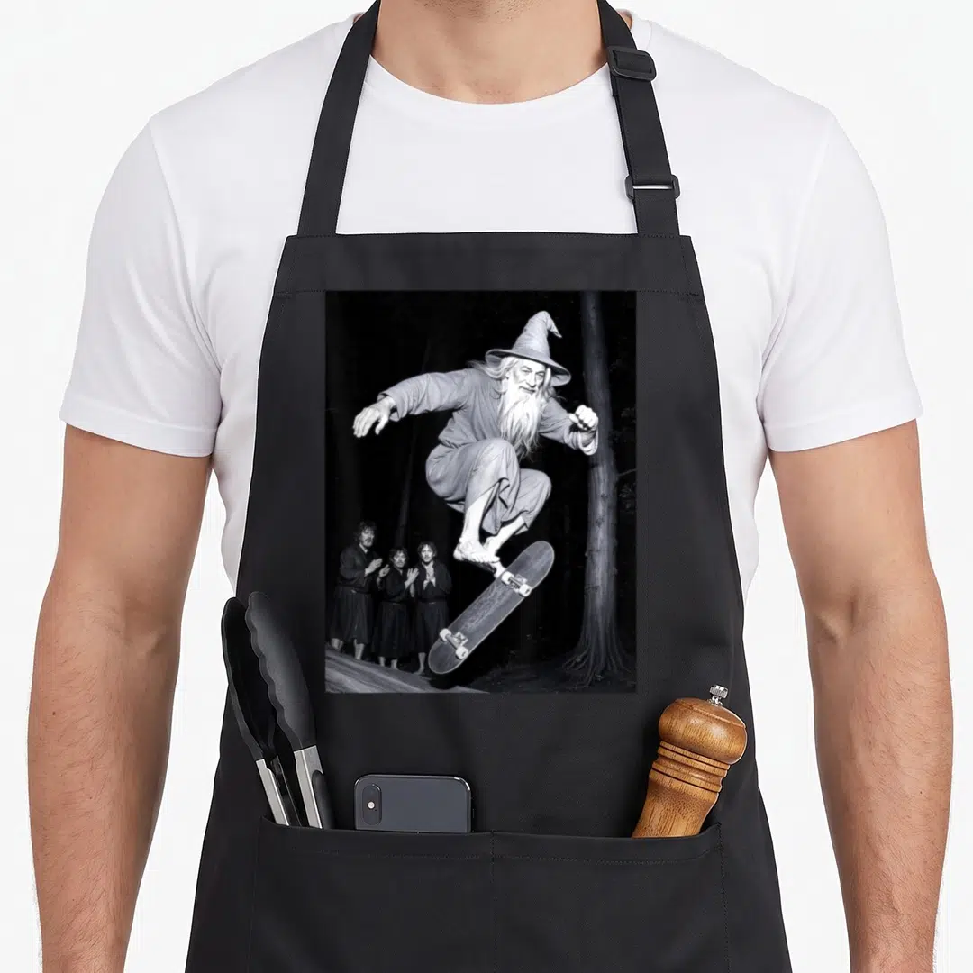 Skateboarding see wizard apron - Wizard jumping on skateboard with three observers in black and white photo — shown on a black cotton canvas apron against a white background