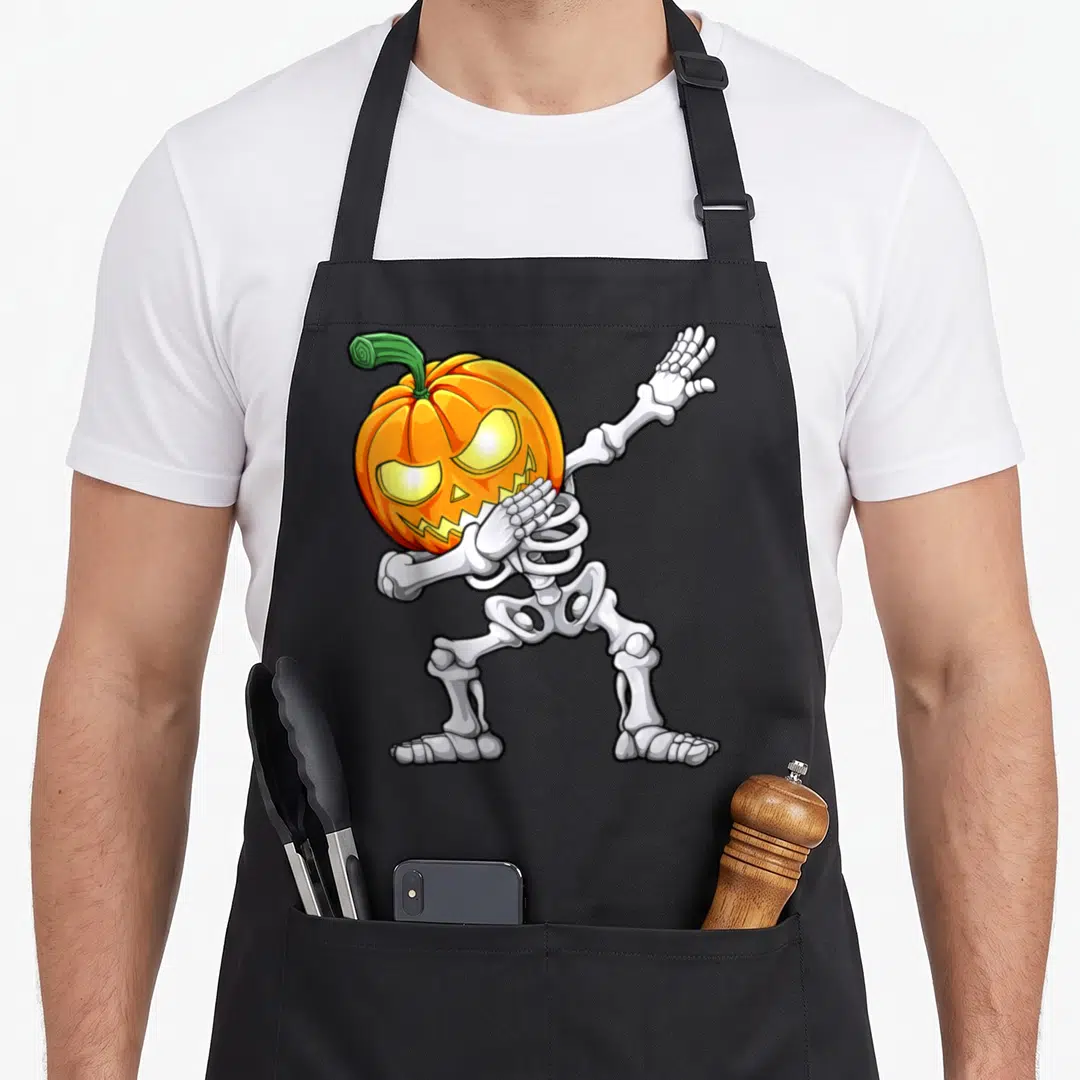 Scary halloween apron - Cartoon skeleton with orange pumpkin head in a dab dance pose with white bones and green stem — shown on a black cotton canvas apron against a white background