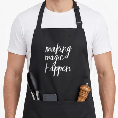 Inspiring making an apron - White stacked script reading making magic happen on grey background — shown on a black cotton canvas apron against a white background