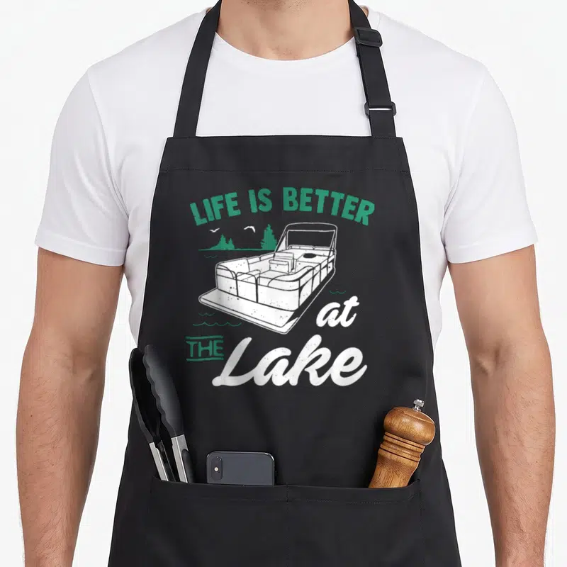 Better boat apron - Retro pontoon boat with LIFE IS BETTER at THE Lake text in green and white — shown on a black cotton canvas apron against a white background