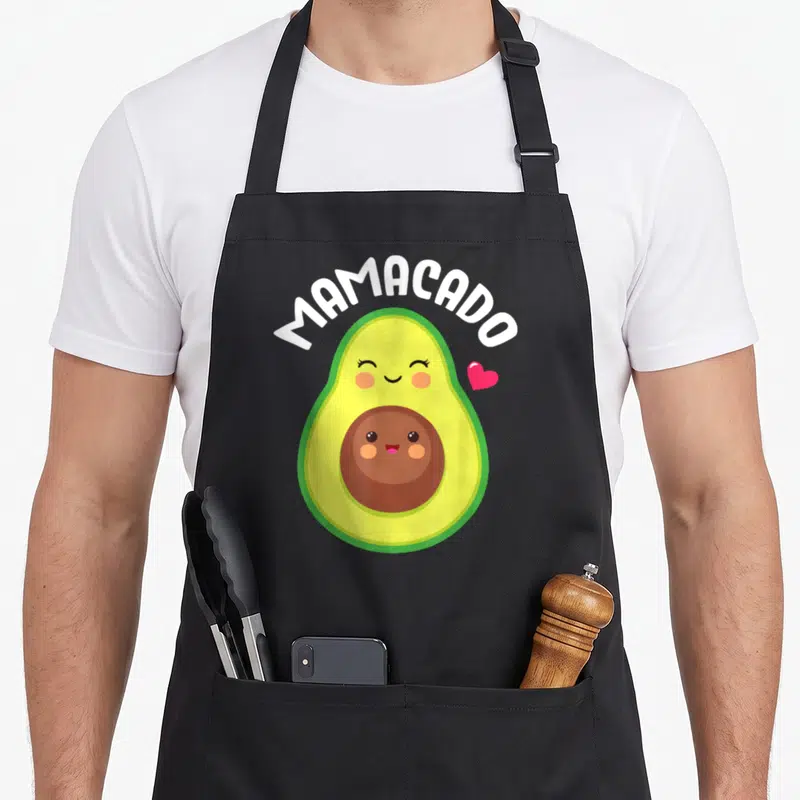 Pregnant avocado apron - MAMACADO text above cartoon avocado with smiling pit and pink heart in green yellow brown — shown on a black cotton canvas apron against a white background