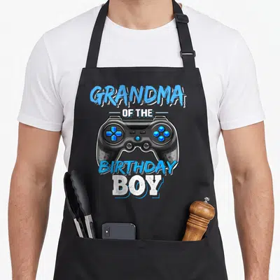 Video grandma in apron - Realistic black game controller with blue buttons and arched text reading GRANDMA — shown on a black cotton canvas apron against a white background