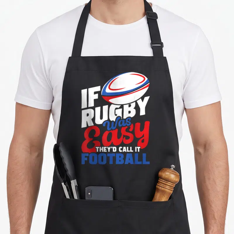 Call easy apron - Rugby ball above stacked text reading IF RUGBY Was Easy THEY'D CALL IT FOOTBALL in white red blue — shown on a black cotton canvas apron against a white background
