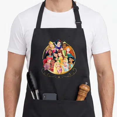 Princess friends cooking apron - Disney Princesses clustered in a gold circle wearing pink blue yellow and gold dresses — shown on a black cotton canvas apron against a white background
