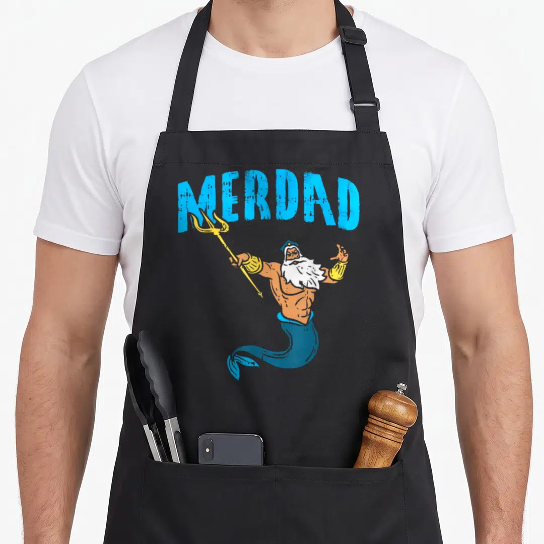 Merdad family aprons - Arched MERDAD text above a cartoon orange merman holding a yellow trident with white beard — shown on a black cotton canvas apron against a white background